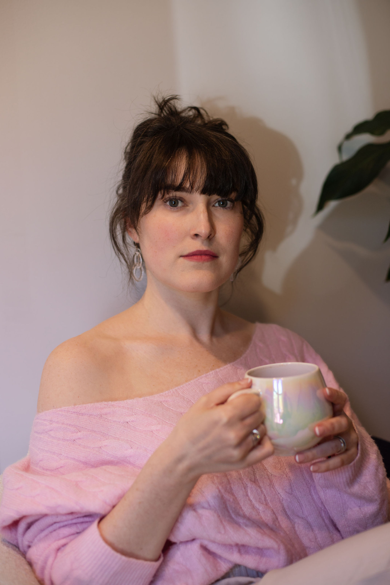 Amélie holding an iridescent mug in an off-shoulder pink knit, a leafy plant to one side.