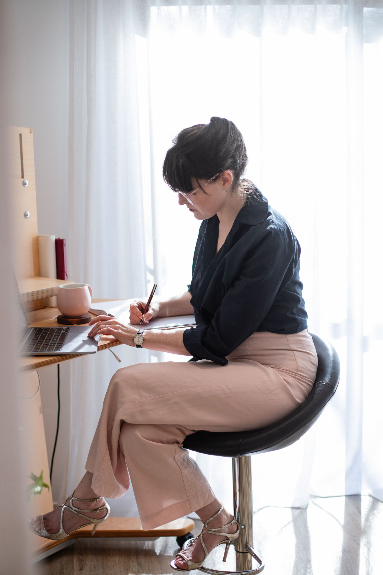 Amélie writing in a notebook at a wall-mounted desk, backlit by a curtained window, a laptop and pink mug beside her.