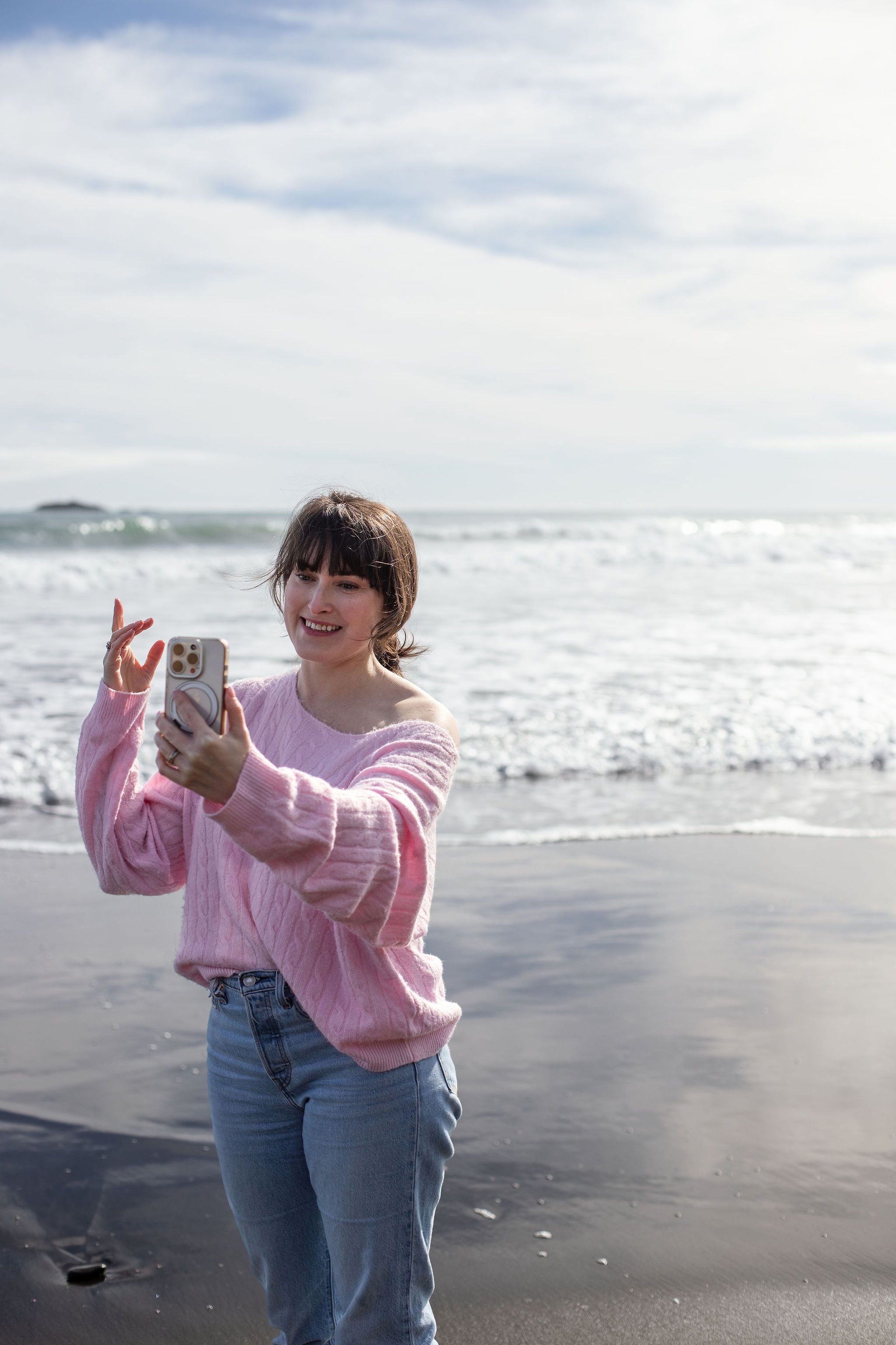 A woman in a pink jumper filming herself on her phone at the beach, smiling into the camera.