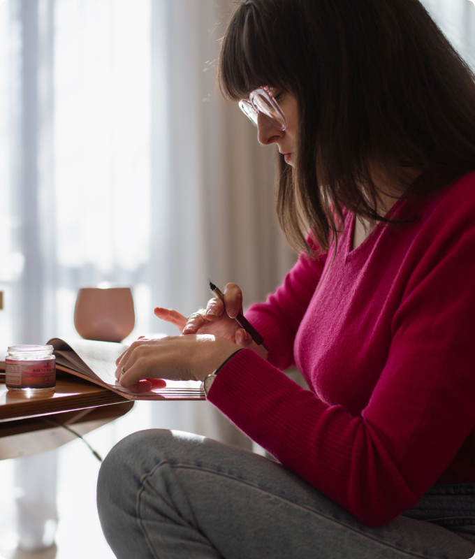 A woman in glasses writing in a notebook by a bright window, a small skincare jar beside her.
