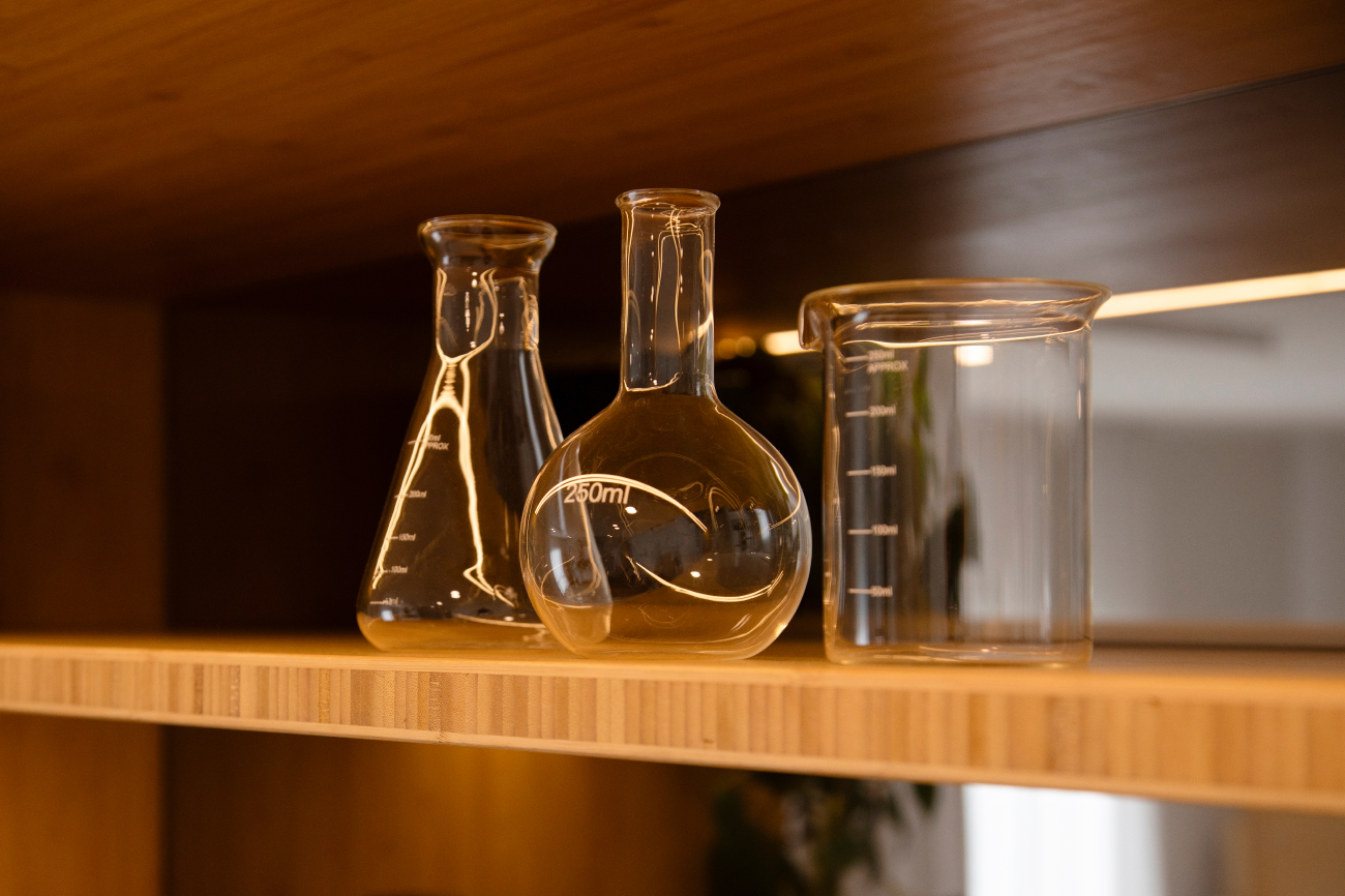 Glass laboratory flasks and beakers arranged on a wooden shelf, softly backlit.