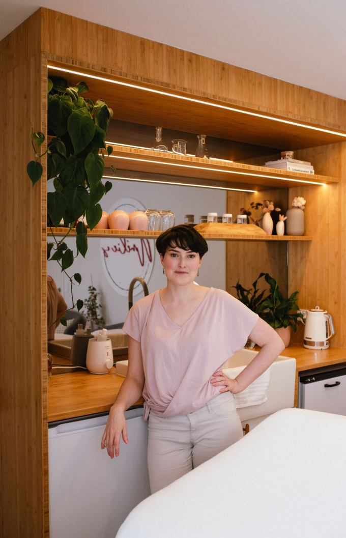 Amélie standing in The Cocoon beside timber shelving styled with plants and skincare.