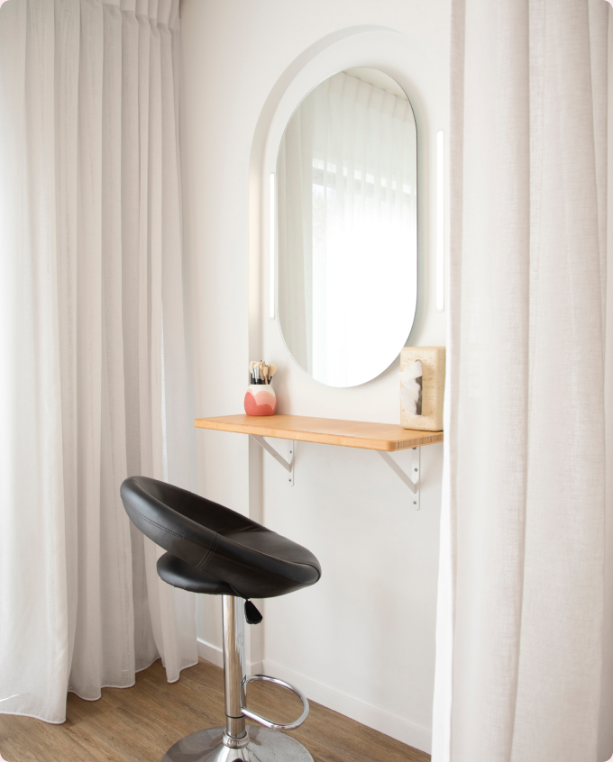 A makeup station with an oval mirror, a black adjustable stool and sheer curtains.