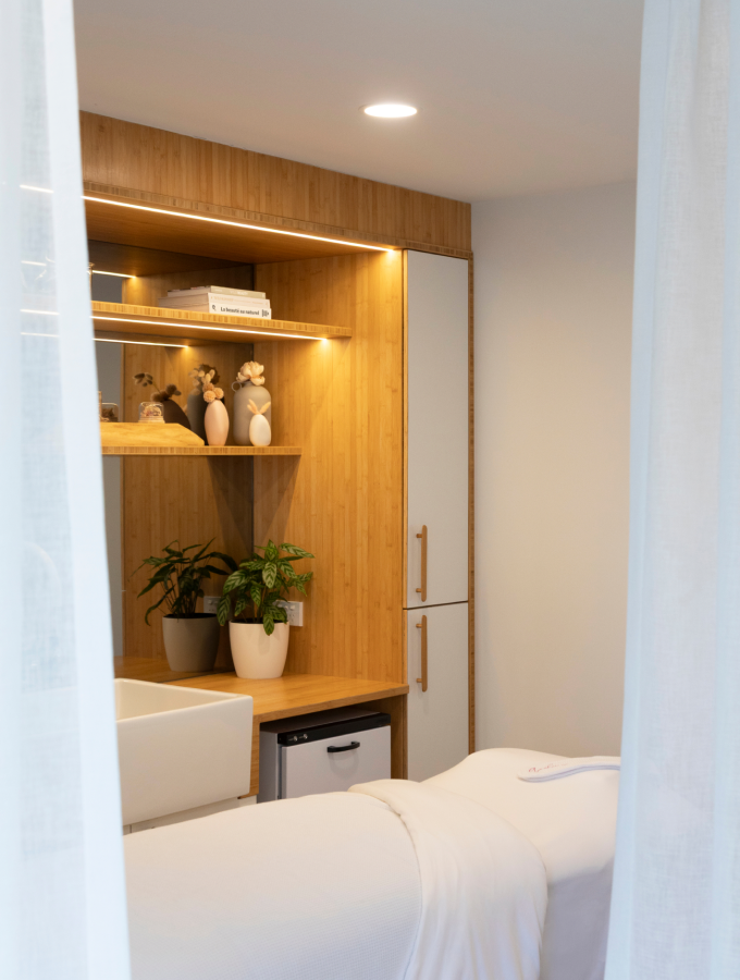 The Cocoon treatment room, softly lit with warm timber shelving and a linen-draped treatment bed behind a sheer curtain.