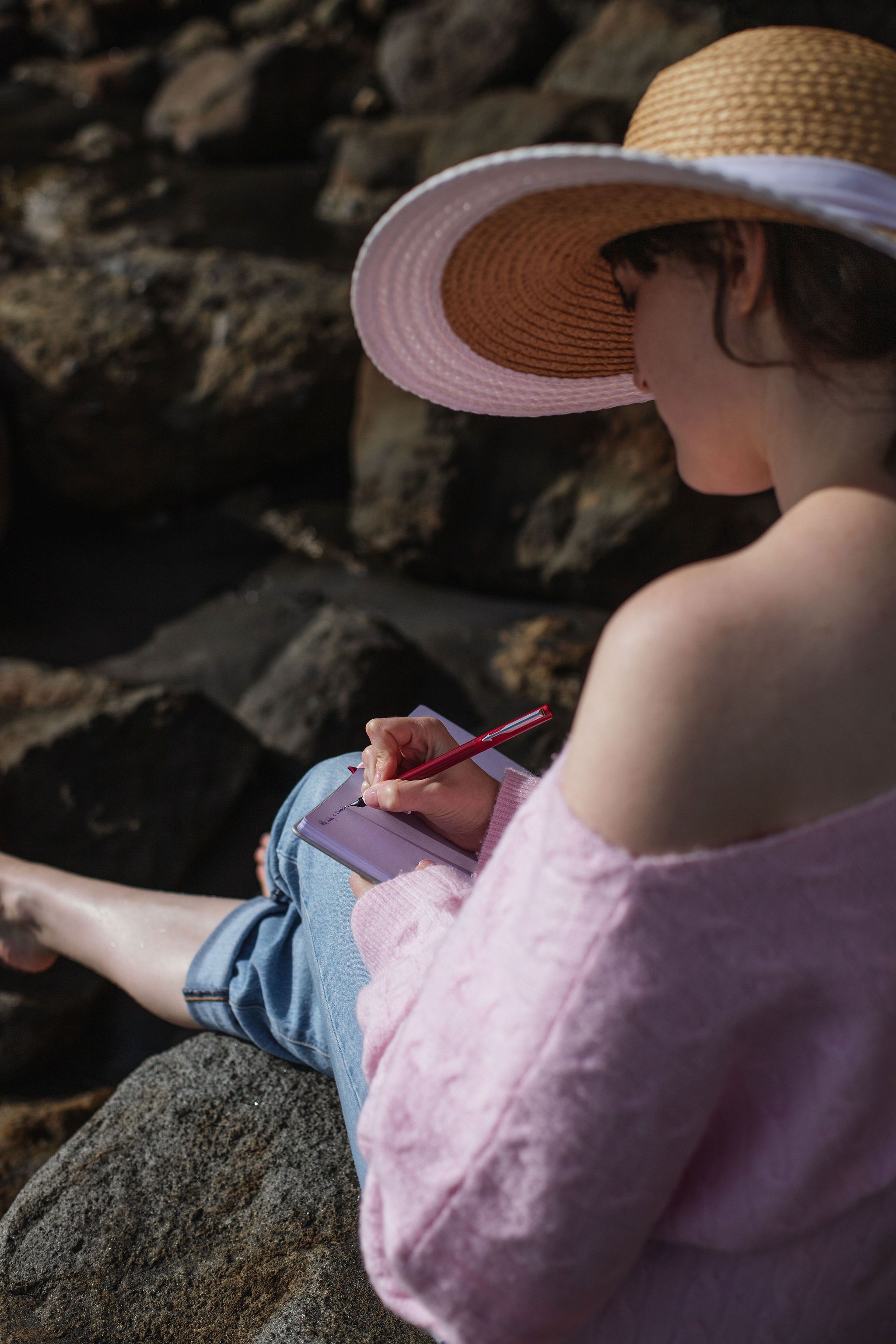 A woman in a sun hat and pink jumper writing in a journal by the shoreline.