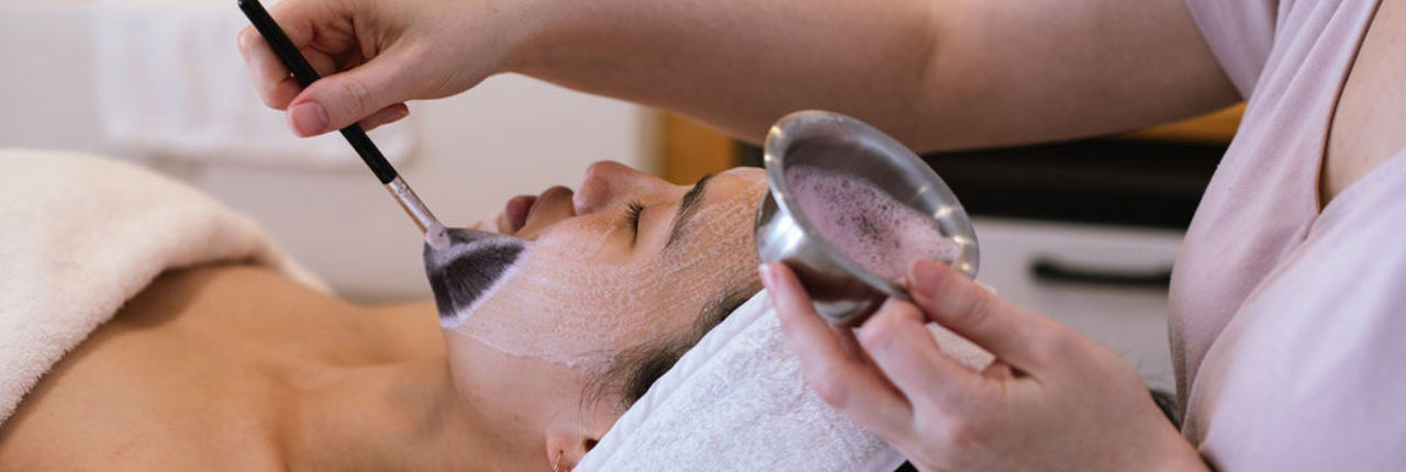 A client resting with a headband while a creamy mask is brushed onto her face during a facial.
