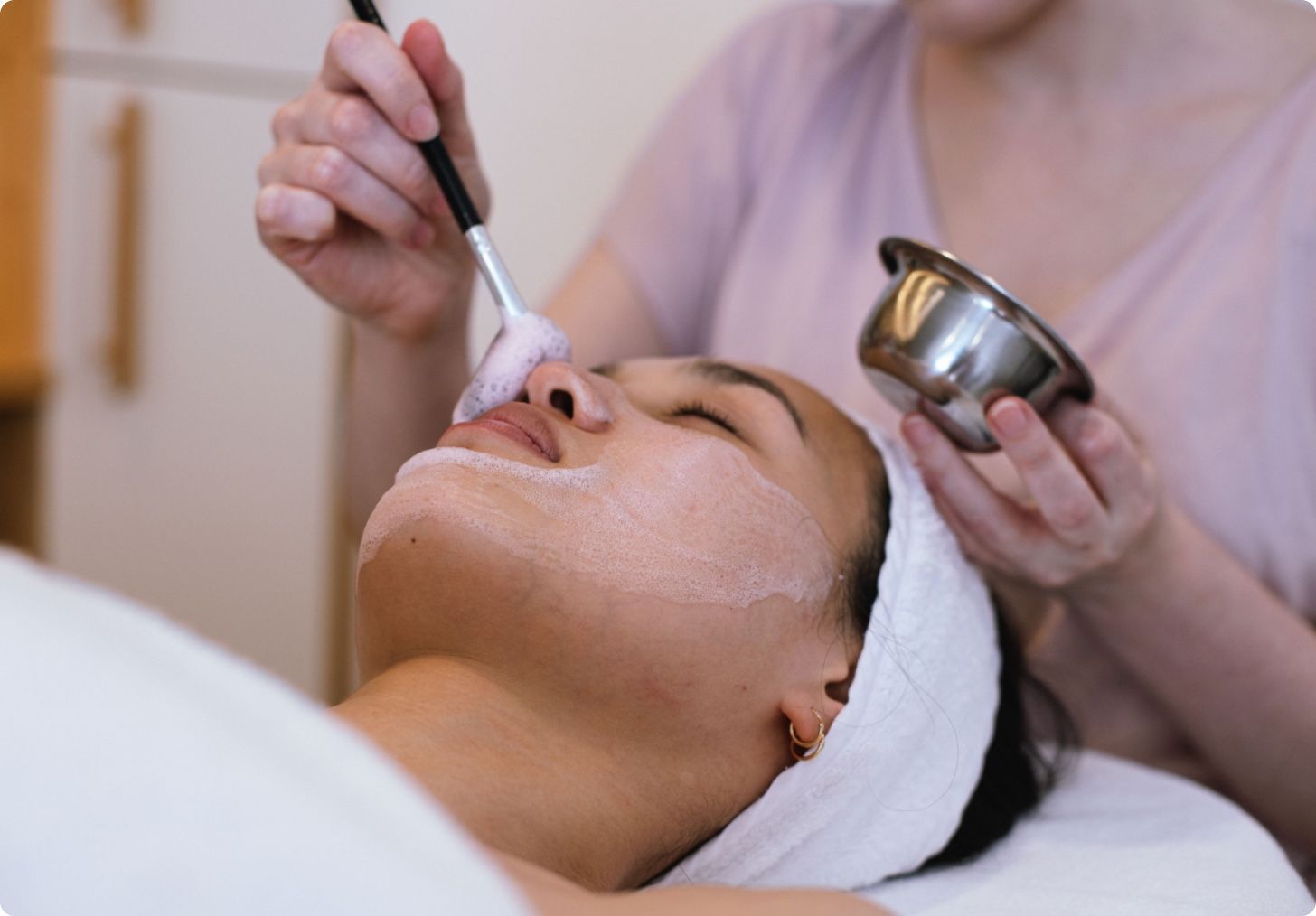 A client resting with a white headband while a creamy mask is brushed onto her face during a facial in the studio.