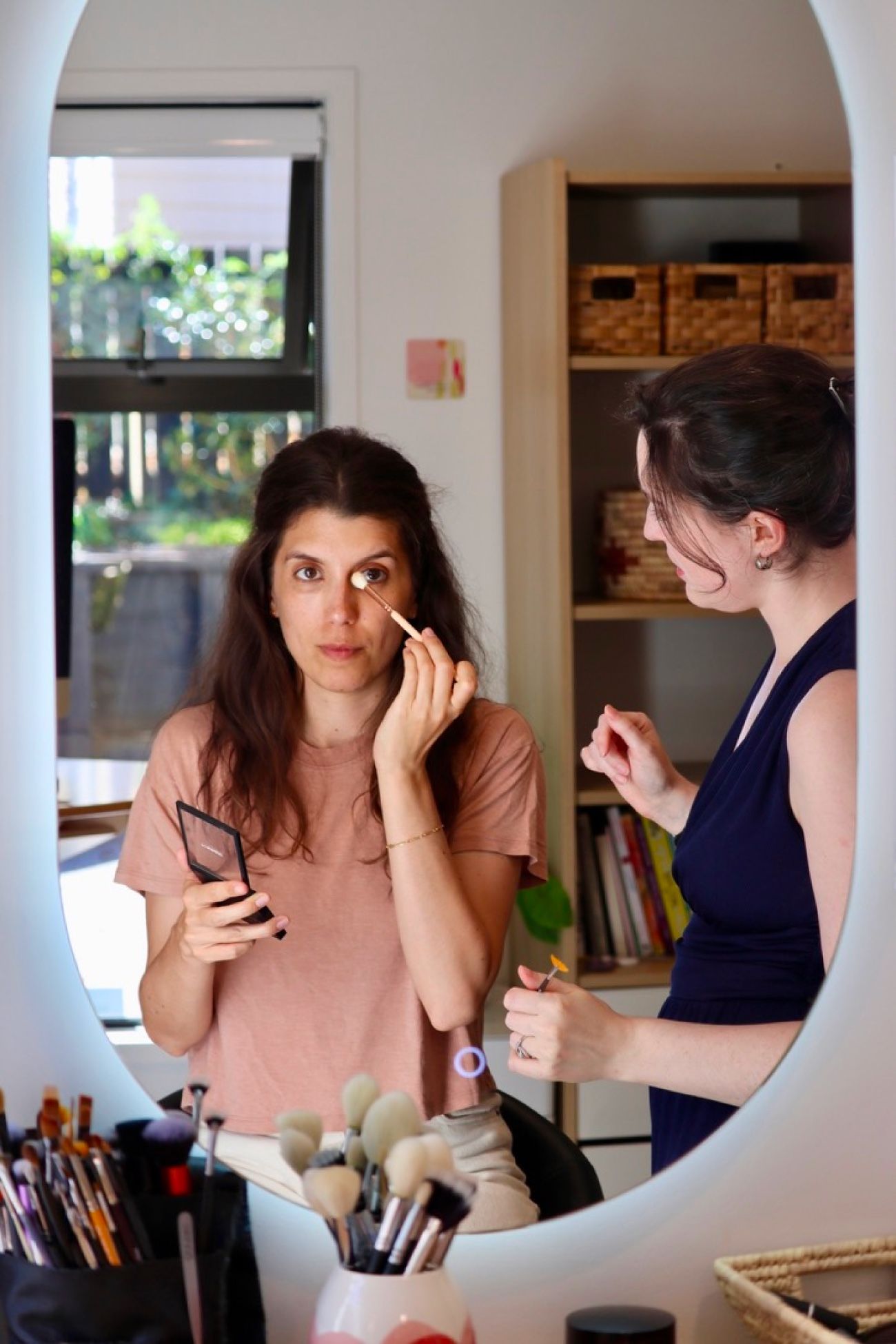 A client applying makeup at the studio's round mirror while Amélie assists, with makeup brushes arranged in the foreground.